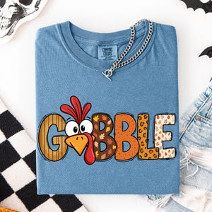 May include: A blue short-sleeve t-shirt featuring the word GOBBLE in large, multi-patterned letters. The letter O is replaced by a cartoon turkey face with a red wattle and orange beak. The other letters feature wood grain, leaf, and leopard prints.