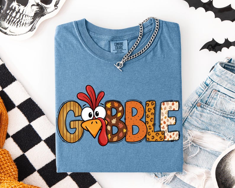 May include: A blue short-sleeve t-shirt featuring the word GOBBLE in large, multi-patterned letters. The letter O is replaced by a cartoon turkey face with a red wattle and orange beak. The other letters feature wood grain, leaf, and leopard prints.