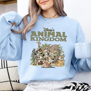 May include: A pale blue sweatshirt with the text "Disney's Animal Kingdom" in green above a cartoon illustration of animals. The design includes a giraffe, zebra, elephant, tiger, and other characters.