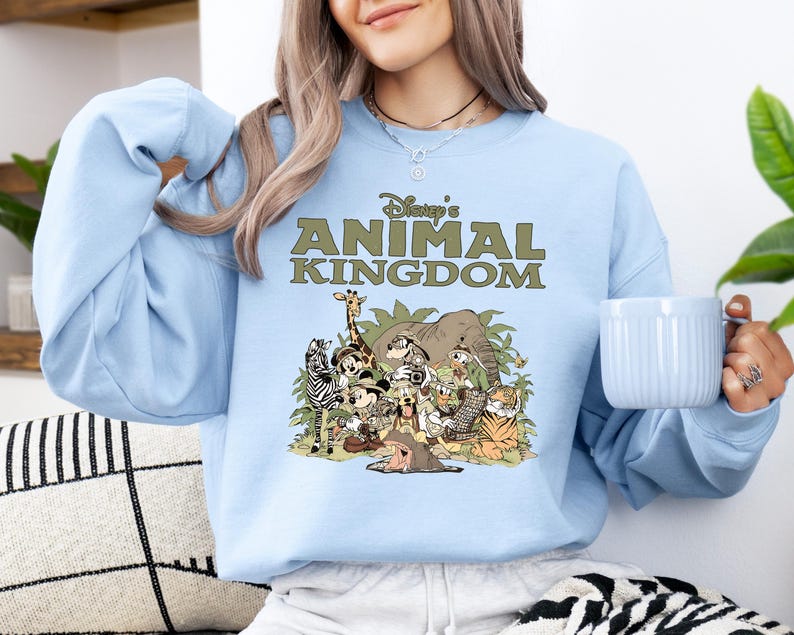 May include: A pale blue sweatshirt with the text "Disney's Animal Kingdom" in green above a cartoon illustration of animals. The design includes a giraffe, zebra, elephant, tiger, and other characters.