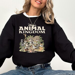 May include: Black sweatshirt with a white collar featuring the text "Disney's Animal Kingdom" in white and tan. The graphic below depicts various Disney characters and animals in a jungle setting. The sweatshirt is a crew neck style.
