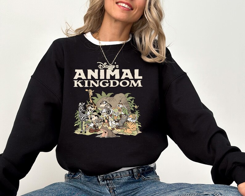 May include: Black sweatshirt with a white collar featuring the text "Disney's Animal Kingdom" in white and tan. The graphic below depicts various Disney characters and animals in a jungle setting. The sweatshirt is a crew neck style.