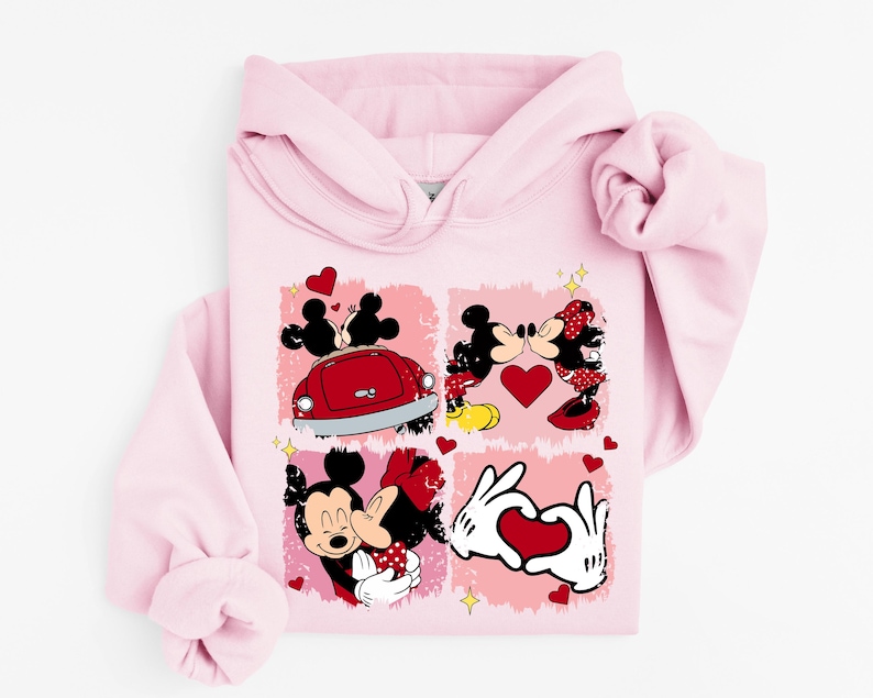 May include: A pale pink hoodie with a graphic of Mickey and Minnie Mouse in romantic scenes. The design includes red hearts and a red car, with Mickey and Minnie kissing and holding hands. The hoodie has a drawstring hood and long sleeves.