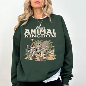 May include: Dark green sweatshirt with the text "Disney's Animal Kingdom" in cream. The graphic below features cartoon animals such as a zebra, giraffe, and tiger. The sweatshirt has a white collar.