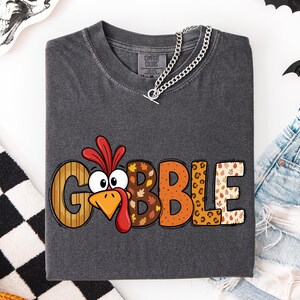 May include: A charcoal gray t-shirt featuring the word GOBBLE printed in large, multicolored letters. The letter O is replaced by a cartoon turkey face with a red wattle. Each letter has a unique pattern including stripes, leaves, and leopard print.
