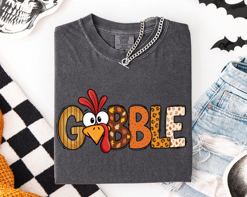 May include: A charcoal gray t-shirt featuring the word GOBBLE printed in large, multicolored letters. The letter O is replaced by a cartoon turkey face with a red wattle. Each letter has a unique pattern including stripes, leaves, and leopard print.