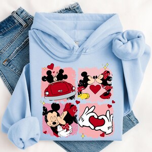 May include: Pale blue hoodie with a graphic of Mickey and Minnie Mouse in romantic scenes. The design includes hearts, a red car, and the characters kissing. The hoodie is folded on top of a pair of blue denim jeans.