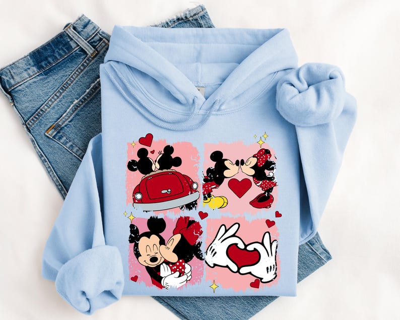 May include: Pale blue hoodie with a graphic of Mickey and Minnie Mouse in romantic scenes. The design includes hearts, a red car, and the characters kissing. The hoodie is folded on top of a pair of blue denim jeans.