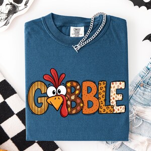 May include: A blue short-sleeve t-shirt featuring the text GOBBLE in large letters. The letter O is replaced by a cartoon turkey face with a red wattle. The letters feature various patterns including wood grain, autumn leaves, and leopard print.
