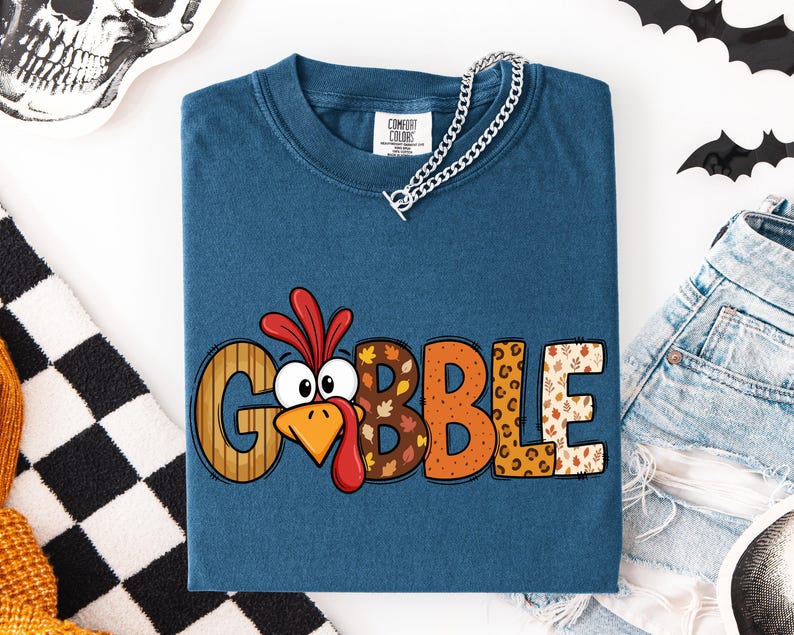 May include: A blue short-sleeve t-shirt featuring the text GOBBLE in large letters. The letter O is replaced by a cartoon turkey face with a red wattle. The letters feature various patterns including wood grain, autumn leaves, and leopard print.