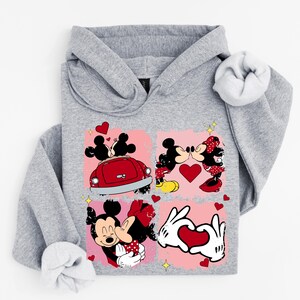 May include: A grey hoodie with a vibrant graphic of Mickey and Minnie Mouse in romantic scenes. The design incorporates hearts, a red car, and the characters kissing. A comfortable and playful design.