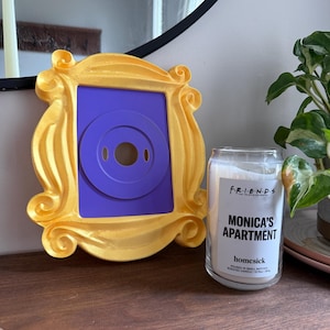May include: A gold-colored picture frame with a purple center, resembling a peephole, sits on a wooden surface. Beside it is a glass jar candle with the text "Monica's Apartment" and "homesick" printed on the label.