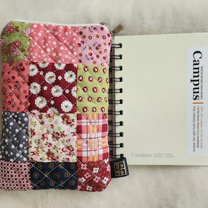 May include: A quilted patchwork pouch with a zipper next to a spiral-bound notebook. The pouch features squares of various floral and dotted fabrics in pink, red, green, and blue. The notebook has the word "Campus" printed on the cover.