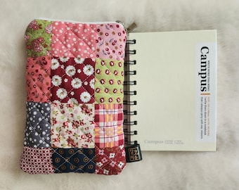 May include: A quilted patchwork pouch with a zipper next to a spiral-bound notebook. The pouch features squares of various floral and dotted fabrics in pink, red, green, and blue. The notebook has the word "Campus" printed on the cover.