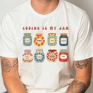 Könnte beinhalten: Weißes T-Shirt mit dem Text "CODING IS MY JAM" in braunen Buchstaben. Das Shirt zeigt Illustrationen von Marmeladengläsern mit Etiketten wie "Debugging" und "Flow". Das Shirt wird von einer Person getragen.