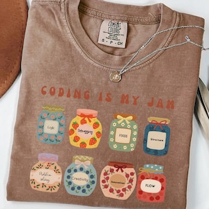 May include: Espresso-colored t-shirt with the phrase "CODING IS MY JAM" and illustrations of jam jars labeled with coding-related terms. A silver necklace with a small pendant is draped across the shirt.