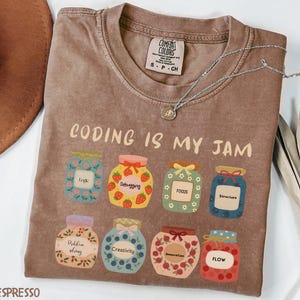 Op de afbeelding: Espresso-kleurig T-shirt met de tekst "CODING IS MY JAM" en geïllustreerde jampotten. Elke pot heeft een ander label, waaronder "Logic", "Debugging" en "Focus". Een zilveren ketting is over het shirt gedrapeerd.