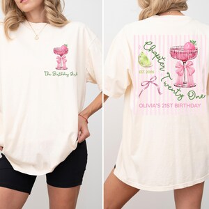 May include: Cream-colored t-shirt with a pink cocktail graphic and the text "The Birthday Girl" on the front. The back features a pink and white striped design with a cocktail, lime, bow, and the text "Chapter Twenty One".