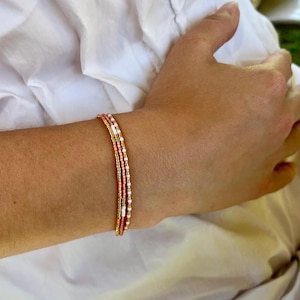 Puede incluir: Un delicado conjunto de pulseras de cuentas. Las pulseras presentan una combinación de cuentas doradas, blancas y color coral. Las pulseras están apiladas en una muñeca, mostrando el color y la textura de las cuentas.