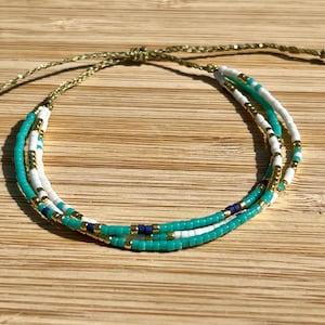 Puede incluir: Una pulsera de cuentas de tres hilos. Presenta cuentas turquesas, blancas y doradas, con algunas cuentas azul oscuro. La pulsera está atada con un cordón dorado.