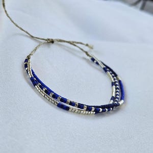 Op de afbeelding: Een armband met meerdere strengen bestaande uit afwisselende segmenten van kleine, diepblauwe en metallic zilveren cilindrische kralen. De strengen zijn verbonden door een dun, lichtbruin natuurvezelkoord dat uitloopt in verstelbare uiteinden.