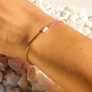 May include: A gold beaded bracelet with a pink and white beaded section. The bracelet is worn on a person's wrist. The background is out of focus, with white rocks.