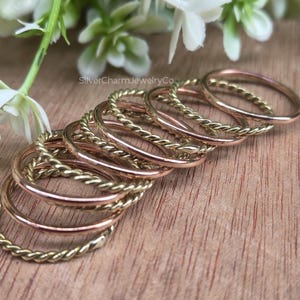 Könnte beinhalten: Eine Sammlung von Ringen in Gold und Roségold, einige mit einem gedrehten Seilmuster, auf einer Holzoberfläche angeordnet. Die Ringe variieren in Stil und Farbe, mit einigen schlichten Bändern. Weiße Blumen und grüne Blätter sind im Hintergrund.