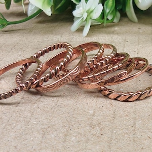 May include: A selection of copper rings with different textures, such as twisted rope and engraved details. The rings are arranged on a neutral surface, with white flowers in the background. The rings have a warm, reddish-brown hue.