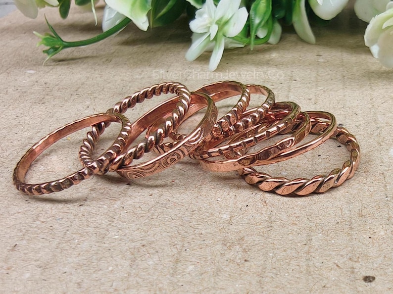 Puede incluir: Un conjunto de anillos de cobre con diversos dise&ntilde;os, incluyendo trenzados y grabados. Los anillos se exhiben sobre una superficie neutra, con flores blancas en el fondo. Los anillos son de un color marr&oacute;n rojizo c&aacute;lido.