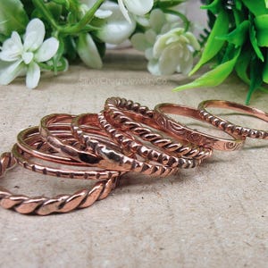 Puede incluir: Una colecci&oacute;n de anillos de color cobre con varios dise&ntilde;os, incluyendo trenzados, con cuentas y grabados. Los anillos est&aacute;n apilados, mostrando sus diferentes texturas y estilos. La imagen est&aacute; sobre un fondo neutro con vegetaci&oacute;n.