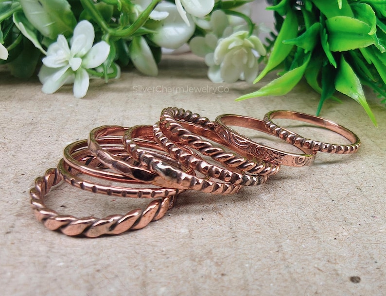 Puede incluir: Una colecci&oacute;n de anillos de color cobre con varios dise&ntilde;os, incluyendo trenzados, con cuentas y grabados. Los anillos est&aacute;n apilados, mostrando sus diferentes texturas y estilos. La imagen est&aacute; sobre un fondo neutro con vegetaci&oacute;n.