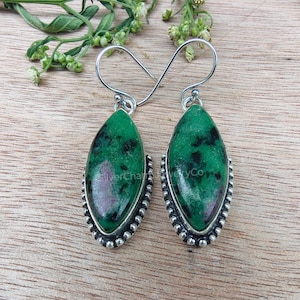 May include: A pair of silver earrings with teardrop-shaped green and pink gemstones. The gemstones are set in a silver bezel with a beaded edge. The earrings are displayed on a wooden surface with greenery.
