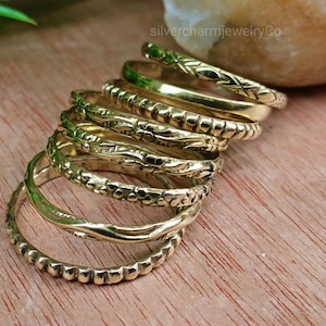 May include: A stack of gold-colored rings with various textured designs, including braided, beaded, and floral patterns. The rings are displayed on a wooden surface, with a natural stone in the background. The rings are likely for fashion or jewelry.