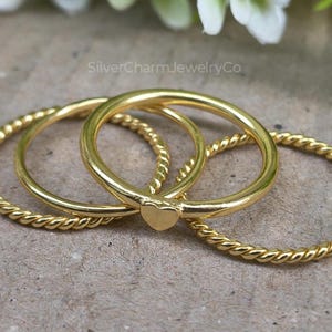 May include: Three gold rings, two with a twisted rope design and one with a small heart accent. The rings are displayed on a neutral surface, with the text "SilverCharmJewelryCo" visible in the background.