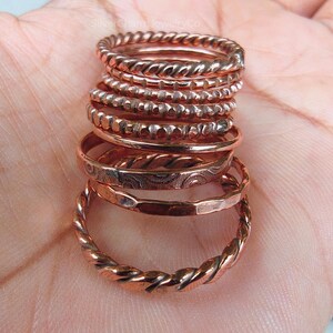 Puede incluir: Una pila de anillos de color cobre con varios dise&ntilde;os, incluyendo trenzados, con cuentas y grabados. Los anillos est&aacute;n apilados, mostrando las diferentes texturas y estilos. Los anillos se exhiben sobre un fondo de color claro.