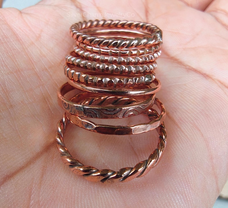 Puede incluir: Una pila de anillos de color cobre con varios dise&ntilde;os, incluyendo trenzados, con cuentas y grabados. Los anillos est&aacute;n apilados, mostrando las diferentes texturas y estilos. Los anillos se exhiben sobre un fondo de color claro.
