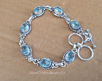May include: A silver bracelet featuring six oval-cut light blue gemstones set in twisted metal frames, connected by silver links and secured with a toggle clasp. The text SilverCharmJewelryCo is visible at the bottom of the image.
