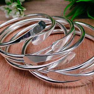 May include: A collection of silver-colored bangles. Each bangle has a smooth, curved design with a flattened, teardrop-shaped accent. The bangles are stacked on a wooden surface, with greenery and white flowers in the background.