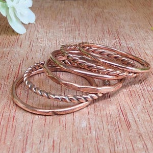 Puede incluir: Un conjunto de anillos de color cobre, algunos con diseños trenzados, dispuestos sobre una superficie de madera. Los anillos varían en estilo y textura, mostrando una mezcla de bandas lisas y texturizadas. Los anillos están apilados, creando un efecto de capas.