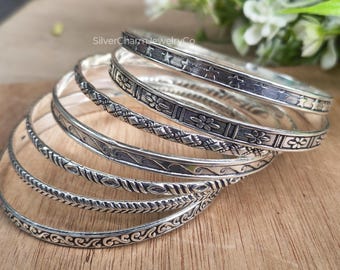 May include: A collection of silver bangles with various embossed designs, including stars, flowers, and geometric patterns. The bangles are stacked on a wooden surface, showcasing their intricate details and textures. The jewelry is from SilverCharmJewelryCo.