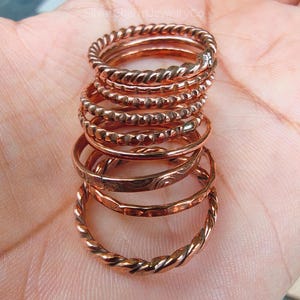 Puede incluir: Una pila de anillos de color cobre con varios dise&ntilde;os, incluyendo trenzados, con cuentas y grabados. Los anillos se muestran sobre un fondo neutro, mostrando sus c&aacute;lidos tonos met&aacute;licos y detalles intrincados. Los anillos son una joya.