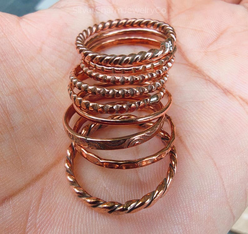Puede incluir: Una pila de anillos de color cobre con varios dise&ntilde;os, incluyendo trenzados, con cuentas y grabados. Los anillos se muestran sobre un fondo neutro, mostrando sus c&aacute;lidos tonos met&aacute;licos y detalles intrincados. Los anillos son una joya.