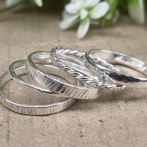 Puede incluir: Un conjunto de anillos de plata con diversas texturas. Uno tiene un diseño de cuerda trenzada, mientras que otros presentan superficies texturizadas o lisas. Los anillos están apilados, mostrando sus diferentes estilos y acabados. Sobre un fondo neutro.