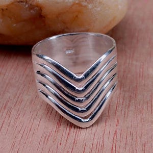 Puede incluir: Un anillo de plata con un diseño de cheurón. El anillo presenta múltiples capas de líneas curvas que forman una forma de V. El anillo está hecho de metal brillante y se muestra sobre una superficie de madera.