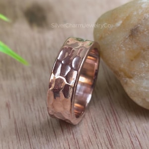 May include: A hammered copper ring with a textured surface. The ring has a wide band and a warm, reddish-brown color. The ring is displayed on a wooden surface.