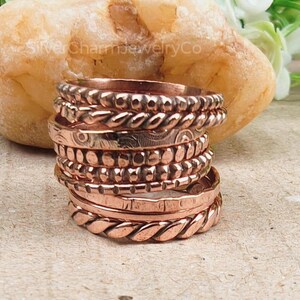 Puede incluir: Una pila de anillos de color cobre con varias texturas, incluyendo dise&ntilde;os trenzados, punteados y grabados. Los anillos est&aacute;n apilados sobre una superficie marr&oacute;n claro, con una gran roca texturizada en el fondo. Los anillos son de un color met&aacute;lico c&aacute;lido.