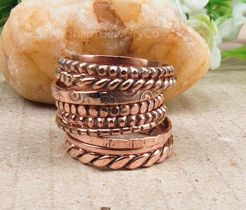 Puede incluir: Una pila de anillos de color cobre con varias texturas, incluyendo dise&ntilde;os trenzados, punteados y grabados. Los anillos est&aacute;n apilados sobre una superficie marr&oacute;n claro, con una gran roca texturizada en el fondo. Los anillos son de un color met&aacute;lico c&aacute;lido.