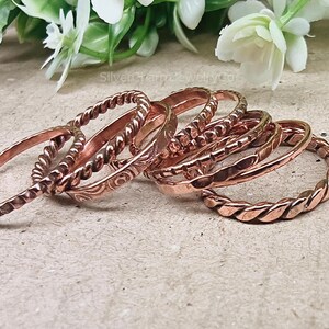 Puede incluir: Una colecci&oacute;n de anillos de color cobre con varios dise&ntilde;os, incluyendo cuerdas trenzadas y patrones grabados. Los anillos est&aacute;n apilados, mostrando sus diferentes texturas y estilos. Las joyas se exhiben sobre una superficie neutra con acentos florales.