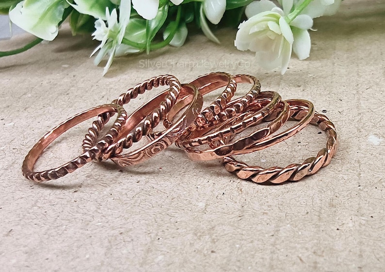 Puede incluir: Una colecci&oacute;n de anillos de color cobre con varios dise&ntilde;os, incluyendo cuerdas trenzadas y patrones grabados. Los anillos est&aacute;n apilados, mostrando sus diferentes texturas y estilos. Las joyas se exhiben sobre una superficie neutra con acentos florales.