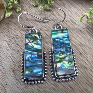 May include: A pair of rectangular earrings with iridescent abalone shell inlays. The earrings have a silver-colored metal frame with a beaded border and a hook closure. The abalone shell displays shades of blue, green, and purple.